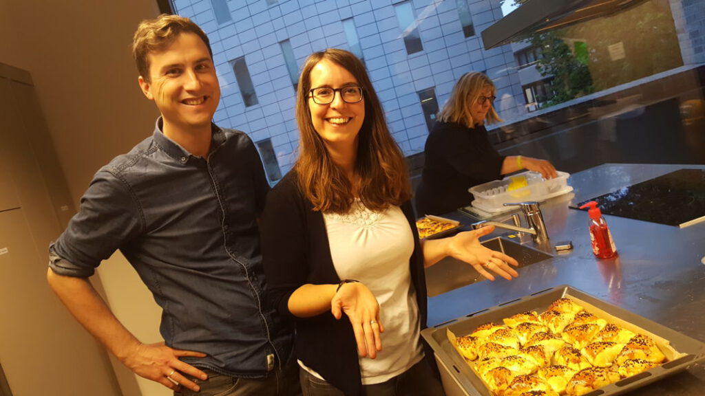Usbekischer Kochkurs 2019 Zwei lächelnde Personen in Küche präsentieren frische, goldbraun gebackene Brötchen auf Backblech, im Hintergrund weitere Person am Spülbecken