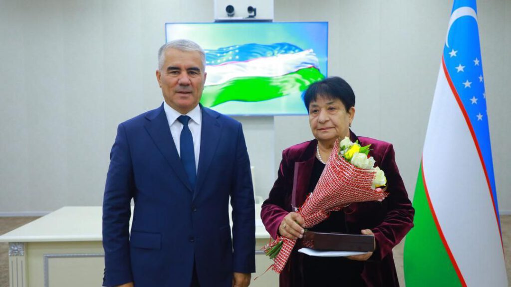 Frau Khushvakht Akramova wurde mit dem Orden „Dustlik“ (Freundschaft) durch Erlass des Präsidenten der Republik Usbekistan ausgezeichnet Zwei Personen bei offizieller Auszeichnung: Mann im dunkelblauen Anzug neben Frau mit Blumenstrauß vor usbekischer Flagge und Bildschirm mit Flaggenmotiv