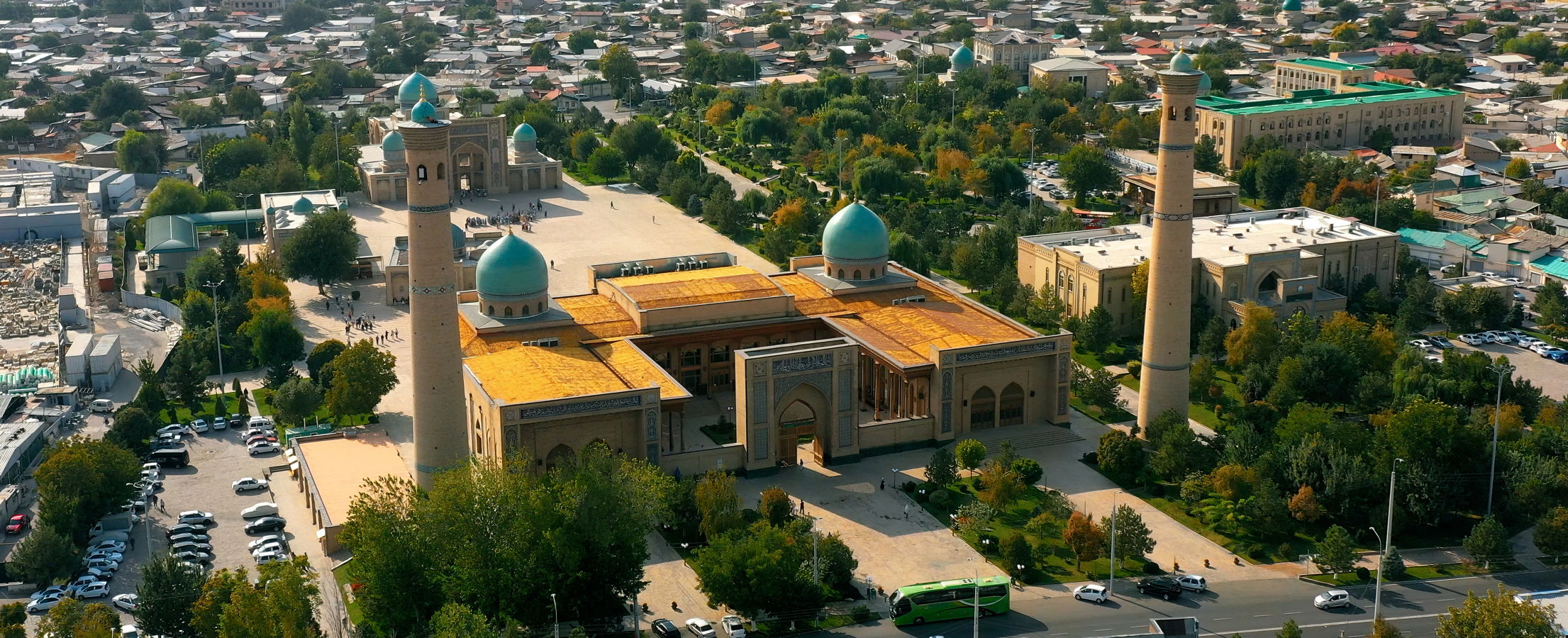 Luftaufnahme einer großen Moschee mit türkisfarbenen Kuppeln, hohen Minaretten und weitläufigem Vorplatz in einer grünen Stadtlandschaft