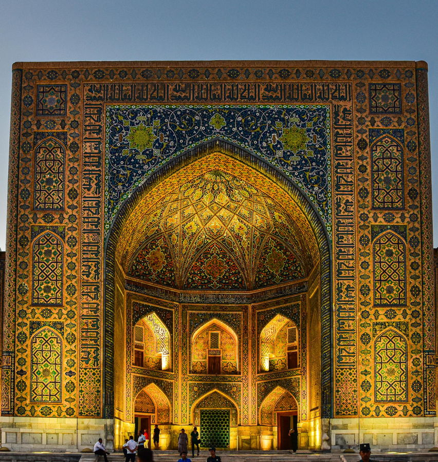 Prächtig beleuchtetes Eingangstor einer islamischen Moschee mit gold-blauen Kachelornamenten, Bögen und detaillierten Mosaiken, Besucher im Vordergrund