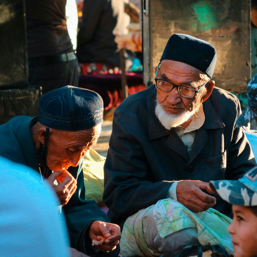 Zwei ältere Männer mit traditioneller Kopfbedeckung und Bart sitzen auf einem Markt, einer isst, der andere trägt Brille und schaut aufmerksam.