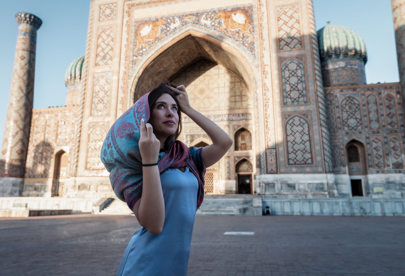 Junge Frau mit buntem Kopftuch vor der aufwendig verzierten Fassade eines zentralasiatischen historischen Medrese-Bauwerks, Reisefotografie